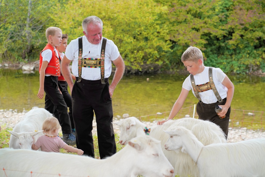 A man and children in traditional dress stand beside white goats; a boy strokes one of the animals.