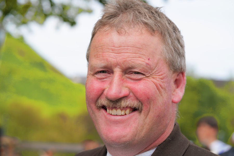 A laughing man with a moustache against a softly blurred green background.
