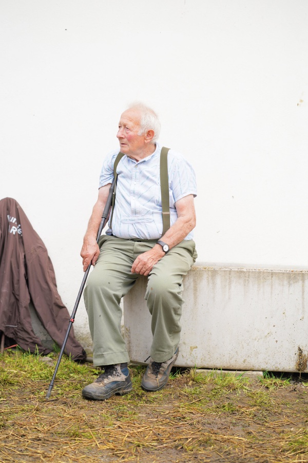 An older man sitting with a walking stick in front of a white wall.