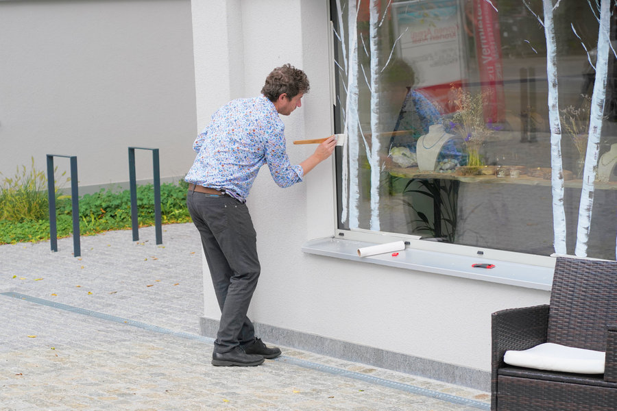 A man bends towards a shop window and works on it with a small tool.