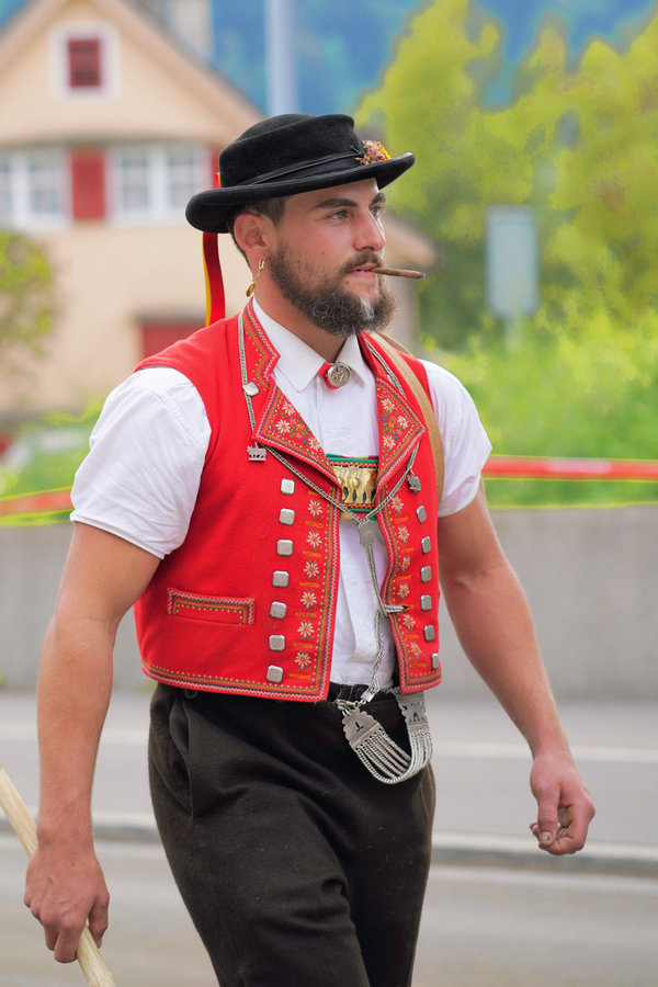 A bearded man in a black hat and red waistcoat in front of village houses.
