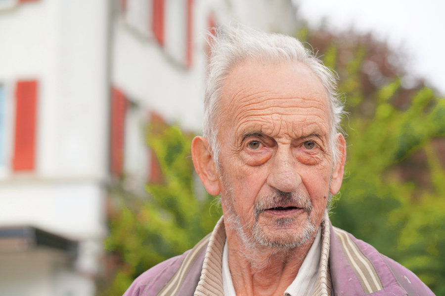 Close-up of a man with grey hair and a beard in front of houses and plants.