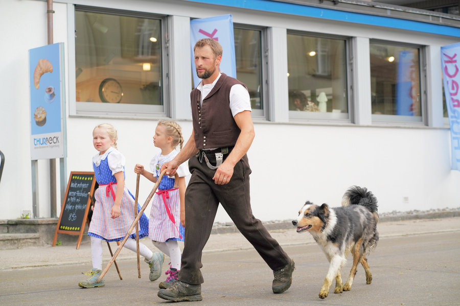Two girls in blue traditional dress beside a man in a dark waistcoat and a tricolour dog.