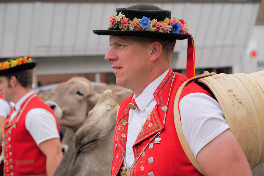 Profile of a man in a flower-decorated hat and red traditional dress, carrying a wooden vessel on his back.