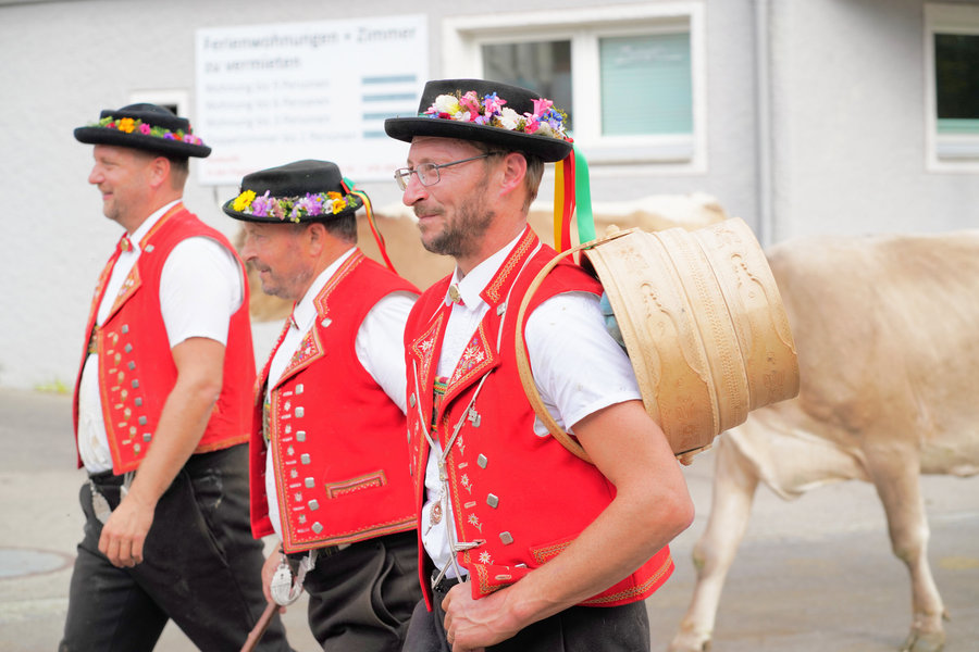 Close side view of three men in red traditional dress, one wearing glasses and carrying a wooden vessel on his back.