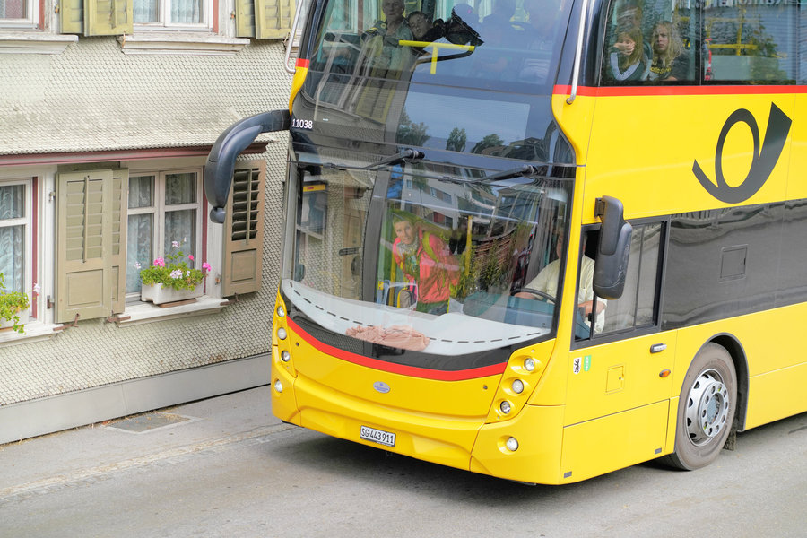 A yellow double-decker PostBus passes an older facade, with passengers visible behind the windscreen.