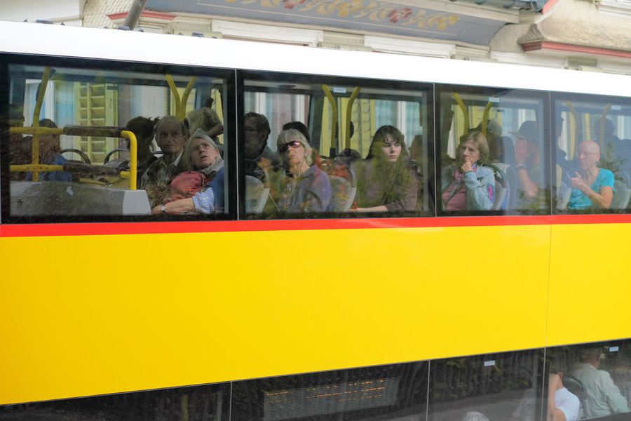 Several passengers look through the upper side windows of a yellow PostBus, some with a hand raised.