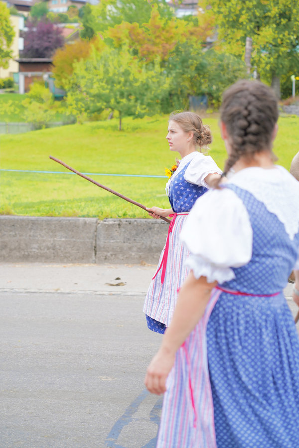 Two women in blue traditional dress carry sticks past a green meadow; one is seen from behind.