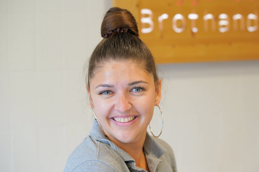 A smiling woman with her hair tied up and wearing a grey top looks towards the camera indoors.