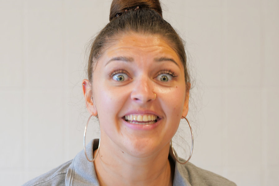 A woman with her hair tied up and wearing a grey top looks towards the camera with a lively expression.