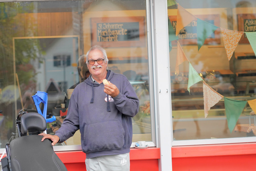 A smiling man wearing glasses holds a piece of pastry beside a chair in front of a shop window.