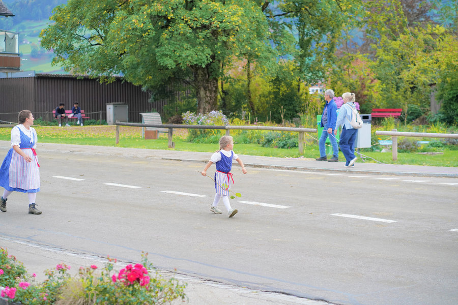 A woman and a girl in blue traditional dress cross the street, with spectators and autumn trees behind them.