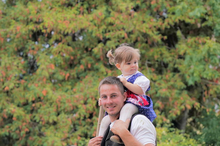 A smiling man carries a little girl in blue traditional dress on his shoulders in front of autumn foliage.