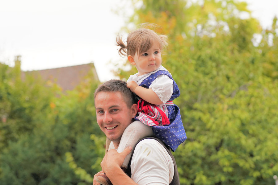 A man carries a little girl in traditional dress on his shoulders while she looks to the side.