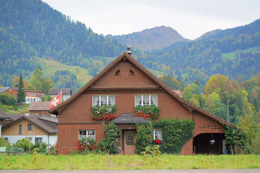 A dark wooden house with red flowers and a Swiss flag in front of green slopes and a rocky mountain.