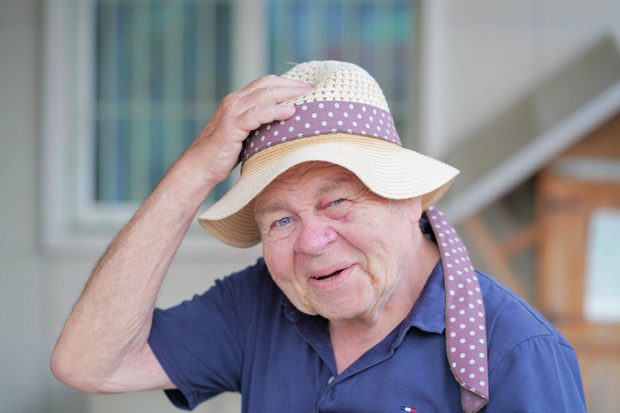 An older man holds his light-coloured hat with a dotted band and looks towards the camera with a lively expression.